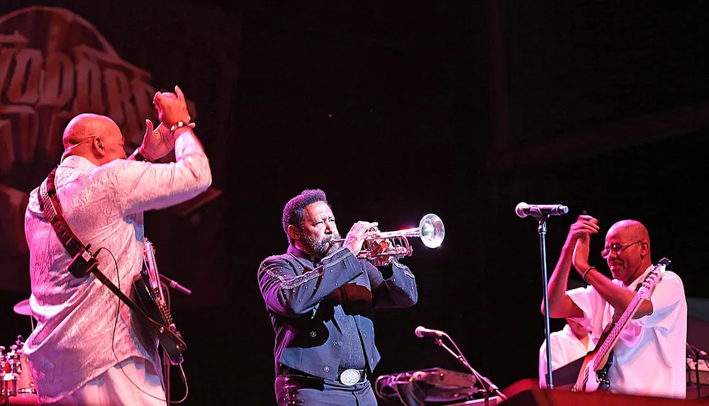 Three musicians are performing on stage with one playing the trumpet another seemingly clapping or engaging with the audience and a third playing an electric bass while sitting down