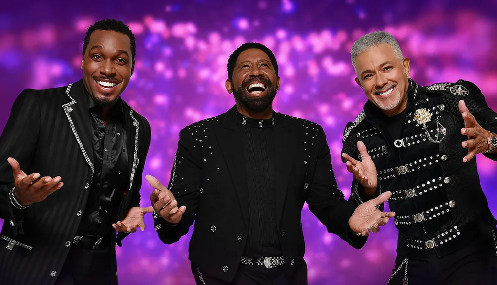 Three cheerful men in embellished black outfits are posing with expressive hand gestures against a vibrant purple bokeh background