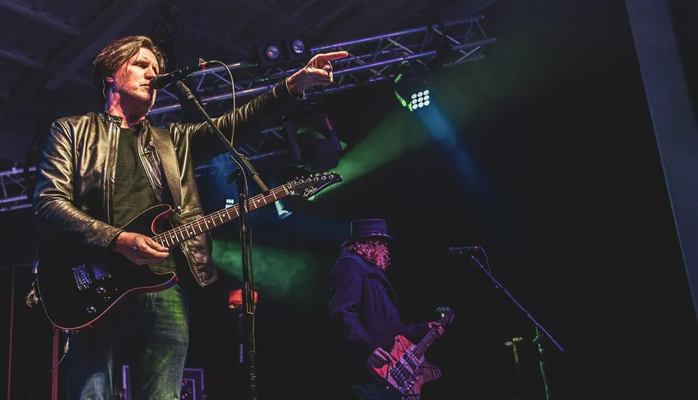 A musician with an electric guitar is performing on stage while pointing out into the audience with another band member in the background playing bass under atmospheric stage lighting