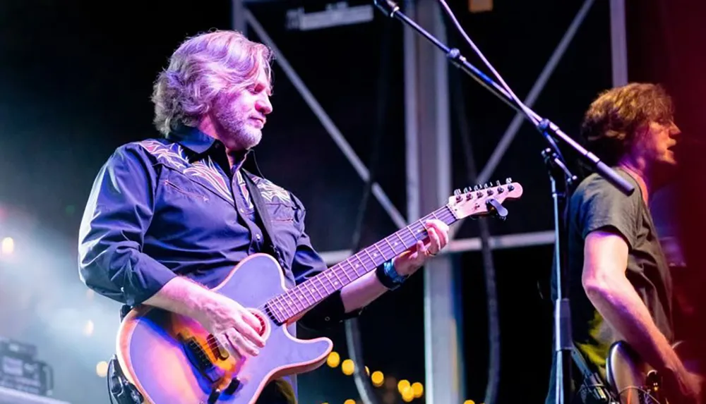 Two musicians are performing on a stage one playing an electric guitar while concentrating on his music