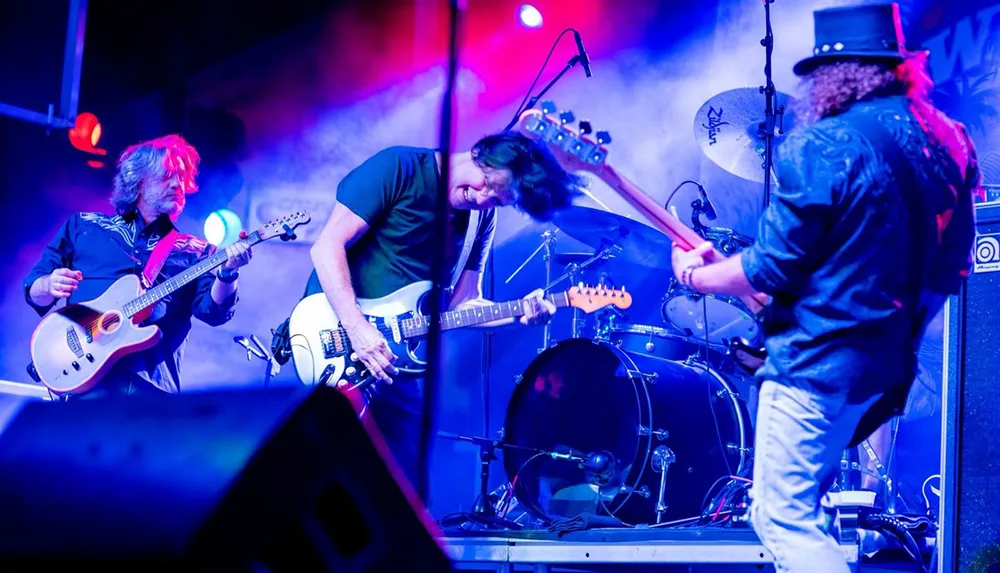 Three musicians are passionately performing in a live concert with two guitarists engaged in their instruments and a drummer in the background amidst vibrant stage lighting