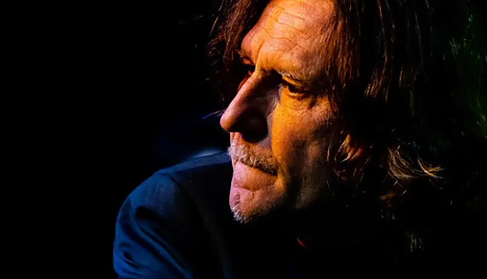 A close-up photo of a man with long hair and a serious expression illuminated by a warm light against a dark background