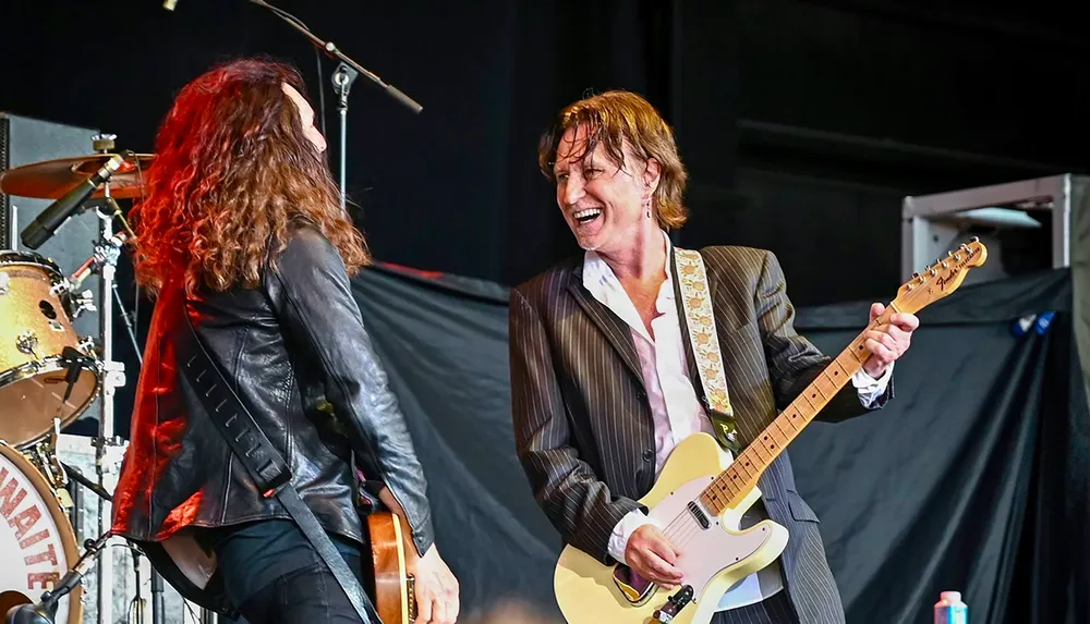 Two musicians are sharing a joyful moment on stage with the man playing a guitar and smiling at his bandmate