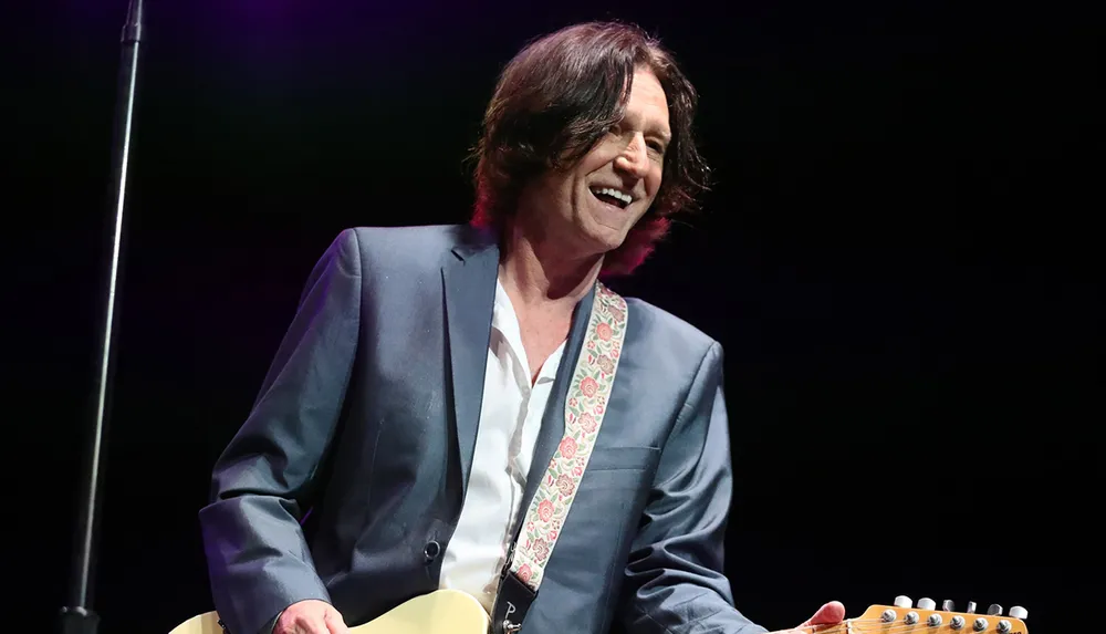 A person with long dark hair smiles while holding a guitar wearing a suit with a floral patterned tie or scarf