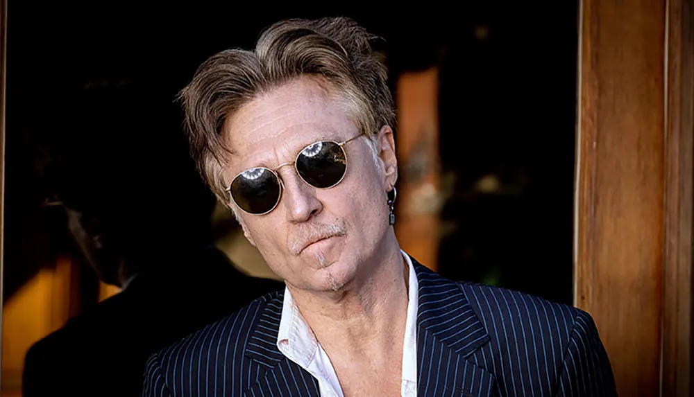 A man with styled hair wearing sunglasses and a pinstriped suit poses confidently