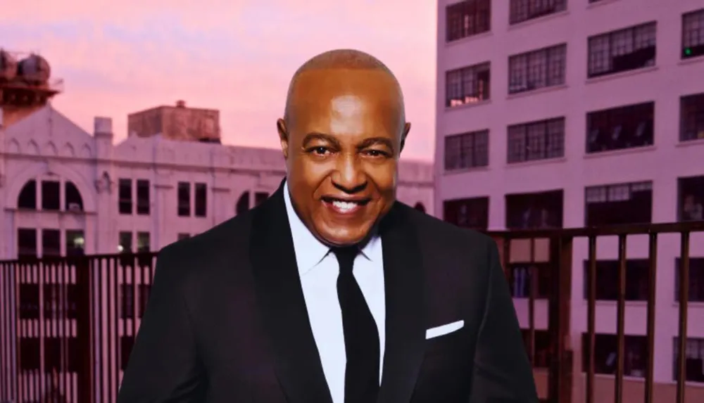 The image shows a smiling person wearing a suit and tie in front of a backdrop that features a building under a sunset sky