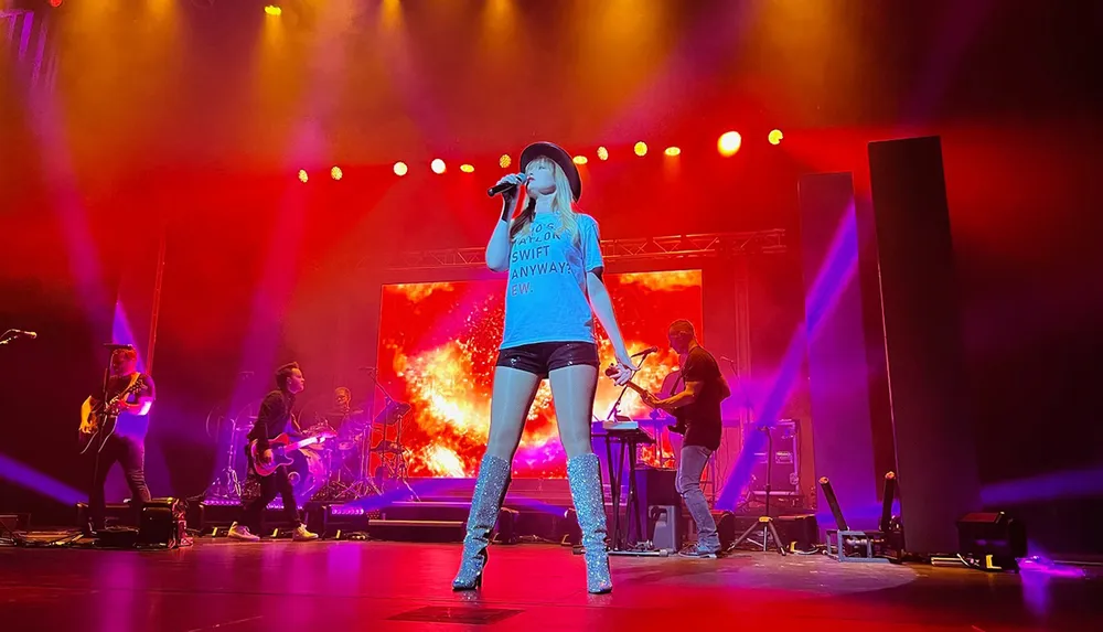 A performer is singing on stage wearing a hat and sparkly thigh-high boots with band members playing instruments in the background amidst vibrant stage lighting