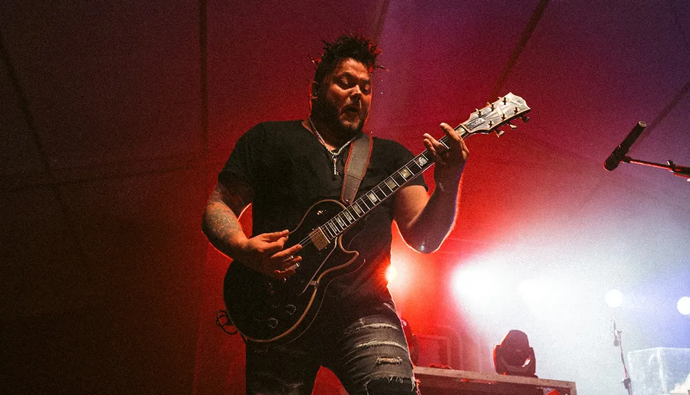 A musician plays an electric guitar on stage immersed in a performance with dramatic stage lighting in the background