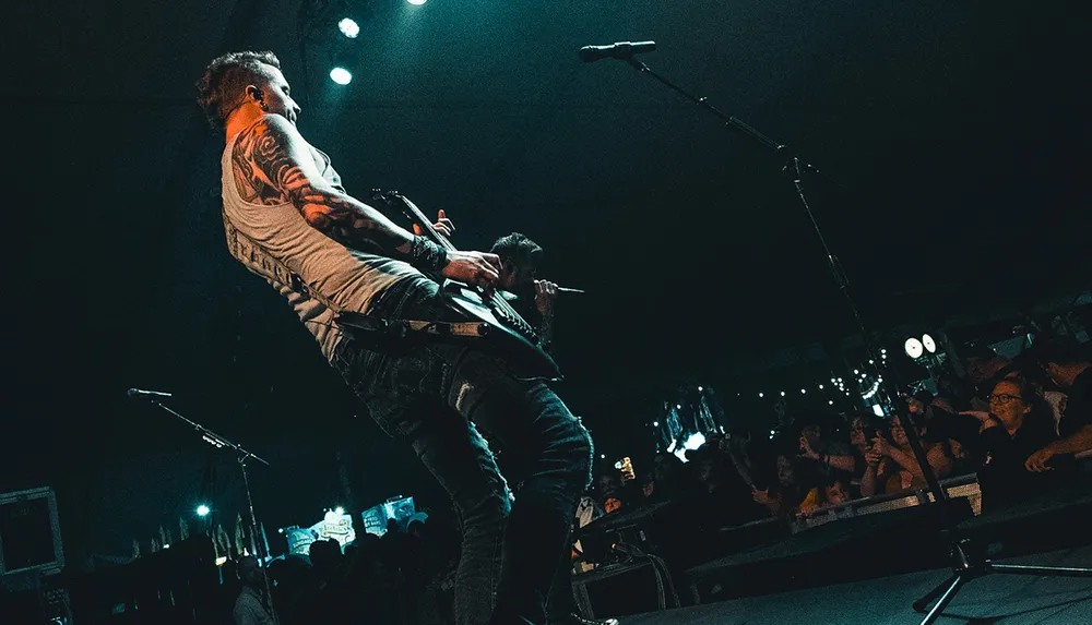 A tattooed guitarist performs energetically on stage in front of an audience