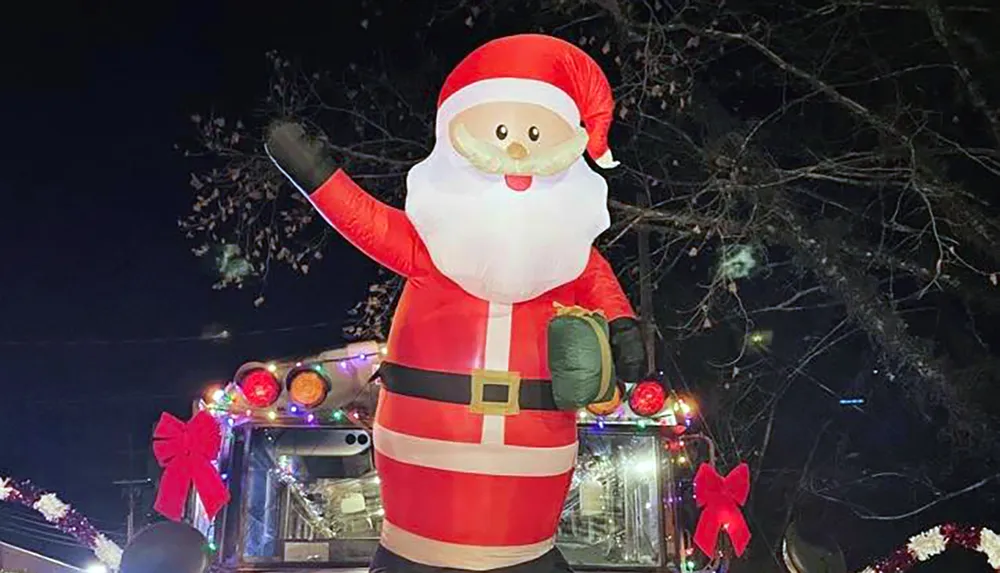 An inflatable Santa Claus decoration is perched atop a vehicle adorned with festive lights and red bows at night