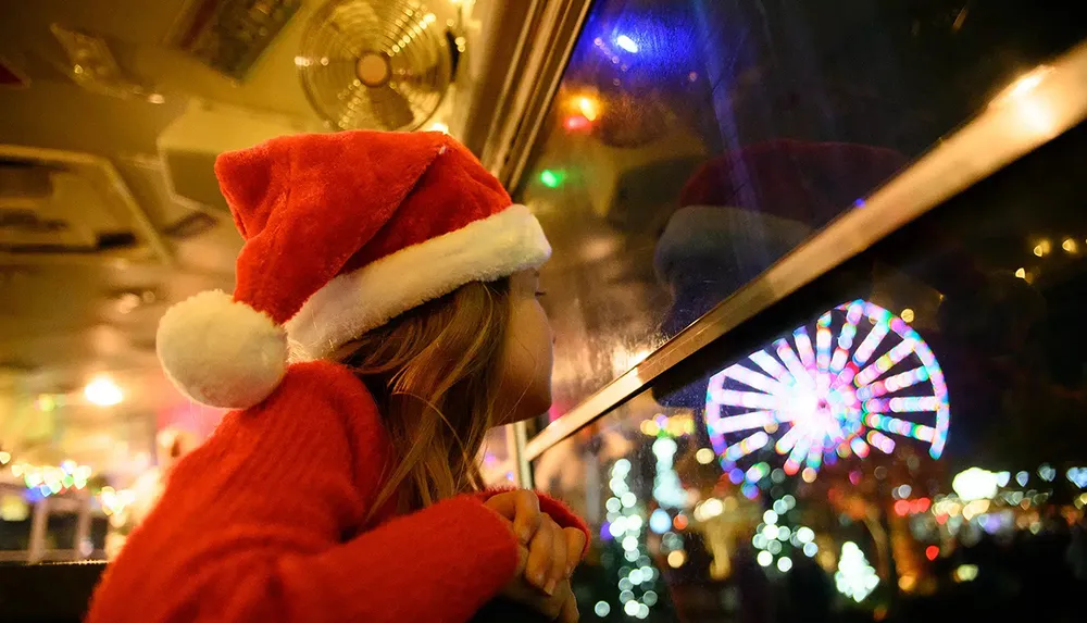 A child in a Santa hat peers longingly out of a window at colorful lights on a festive night