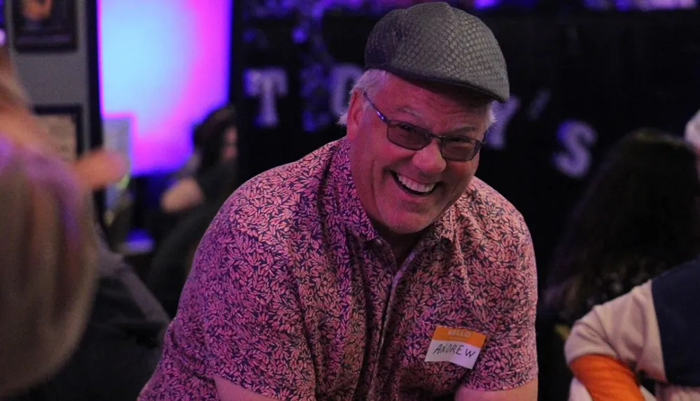 A smiling person in glasses and a patterned shirt is wearing a cap and a name tag seeming to enjoy a social event