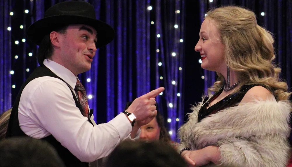 A man in a hat and waistcoat points and smiles at a woman wearing a fur shawl and a beaded dress both seemingly engaged in a jovial moment at a festive event with a blue light backdrop