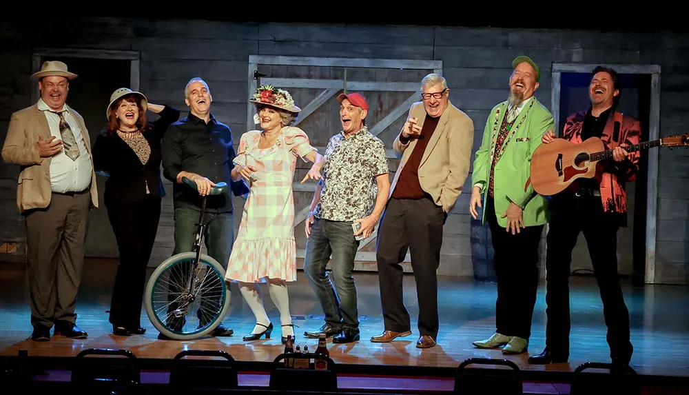 A group of eight people in colorful attire is joyfully posing on a stage with one person holding a guitar and another with a unicycle