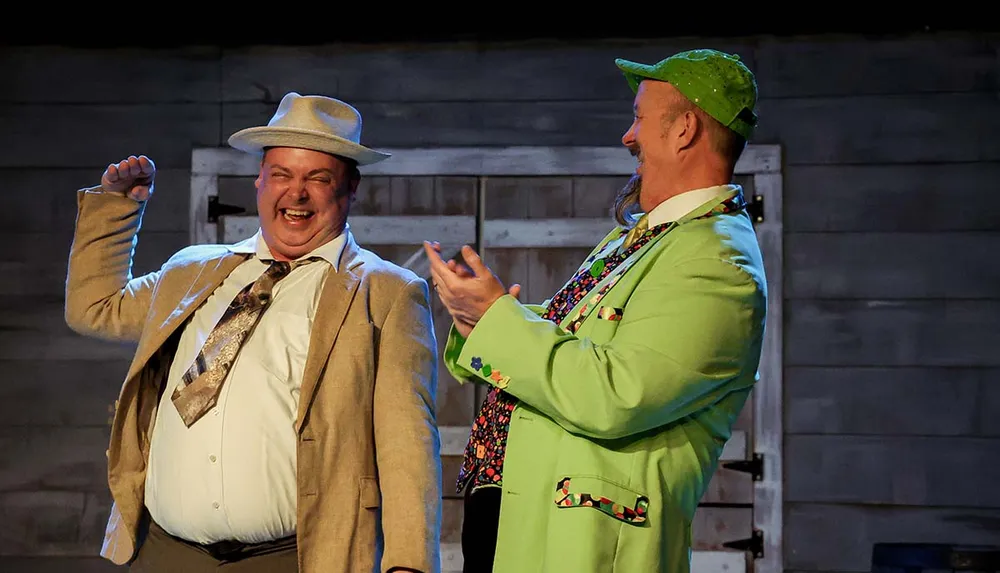 Two men in colorful suits and hats are joyfully laughing and interacting on stage