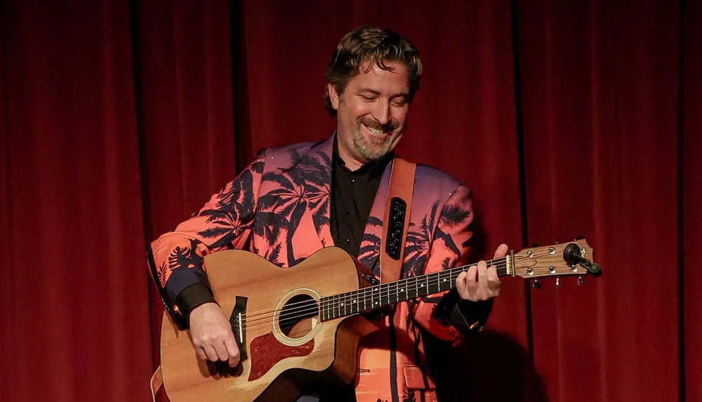 A man in a colorful jacket is playing an acoustic guitar with a smile on his face against a red curtain backdrop