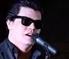 A person wearing sunglasses and a dark suit is singing into a microphone