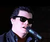 A person wearing sunglasses and a dark suit is singing into a microphone