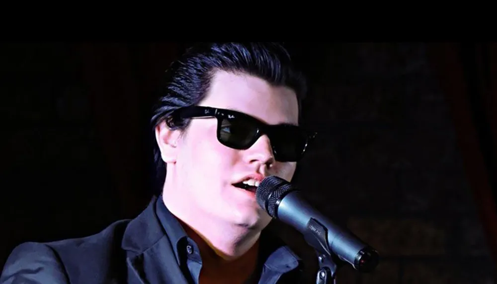 A person wearing sunglasses and a dark suit is singing into a microphone