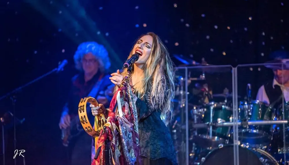 A singer passionately performs on stage holding a tambourine with band members playing instruments in the background