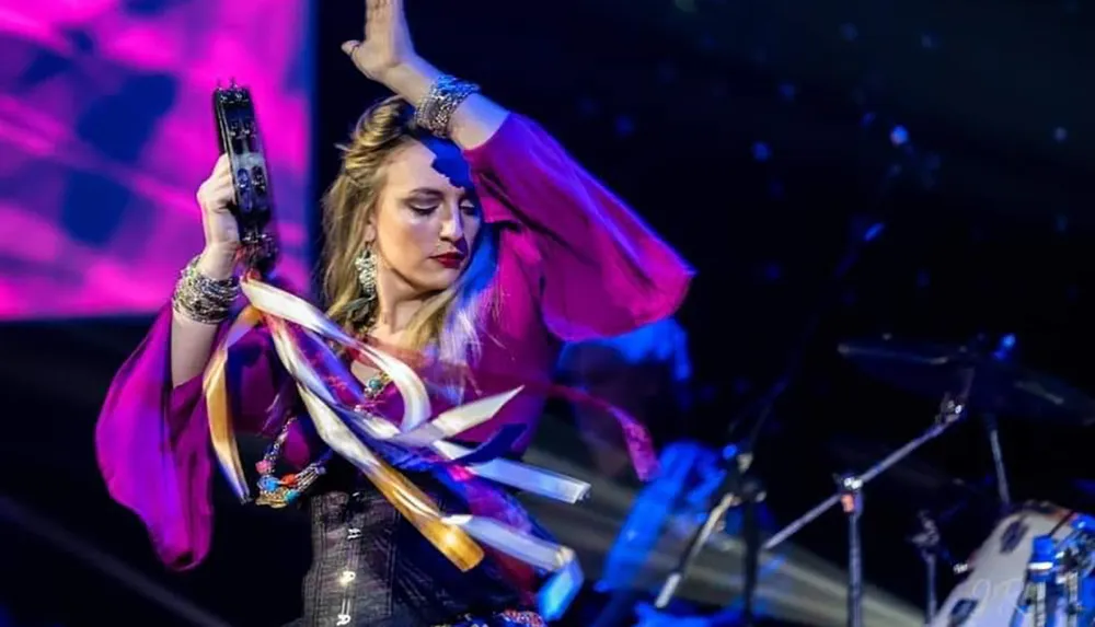 A woman in a vibrant outfit performs passionately on stage with a tambourine set against a colorful and dynamic background