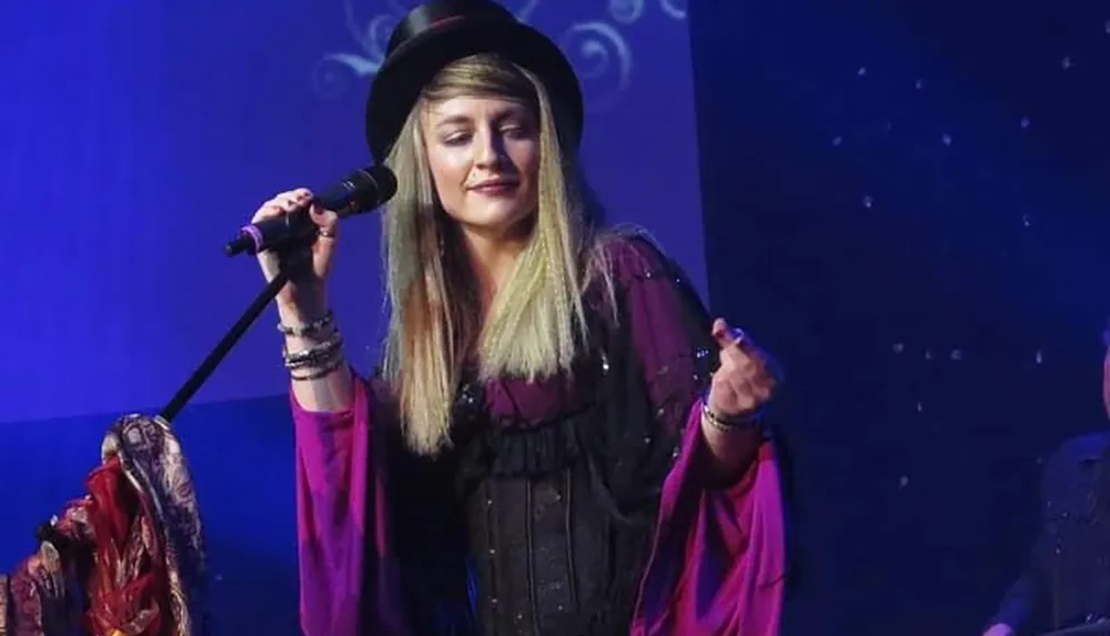 A woman in a hat and purple attire is singing on stage with a microphone