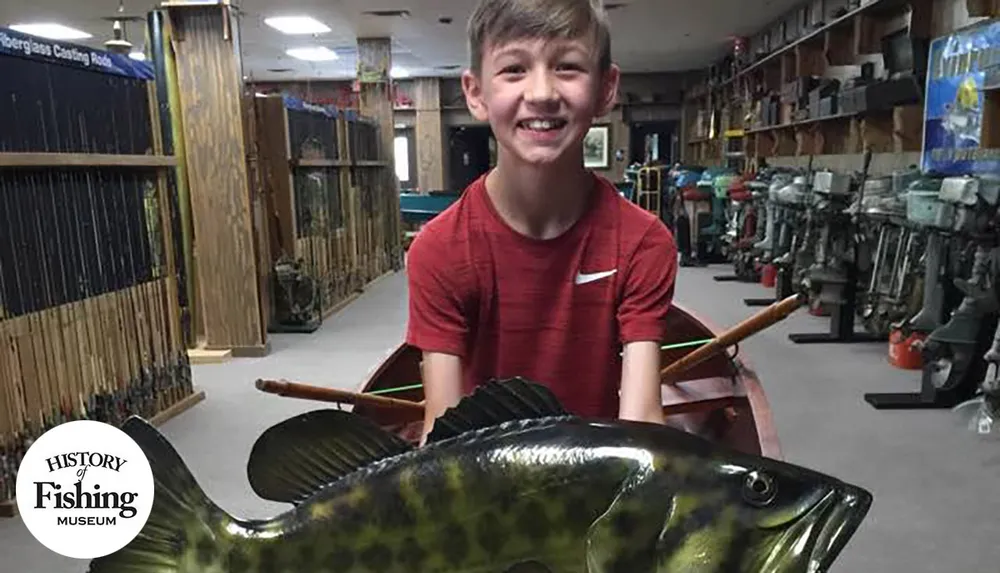 A smiling boy in a red shirt is holding a large model fish inside a fishing museum