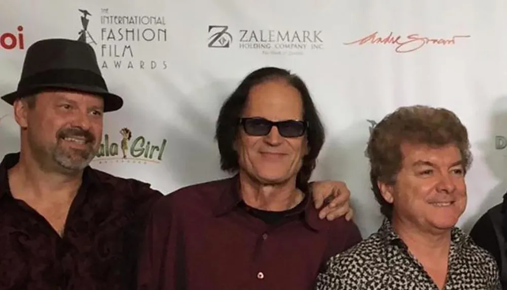 Three men are posing together in front of a backdrop with logos possibly at a film or fashion event