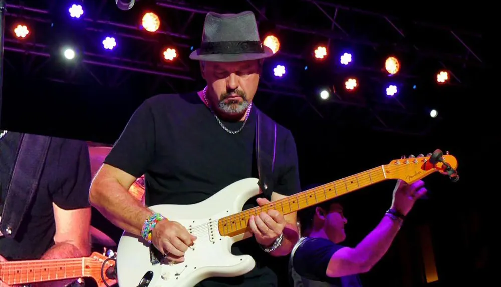 A musician wearing a fedora is playing an electric guitar on stage under colorful lights