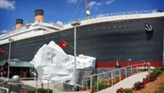 A group of people is observing a large ship model with an iceberg and water feature in a landscaped garden setting.