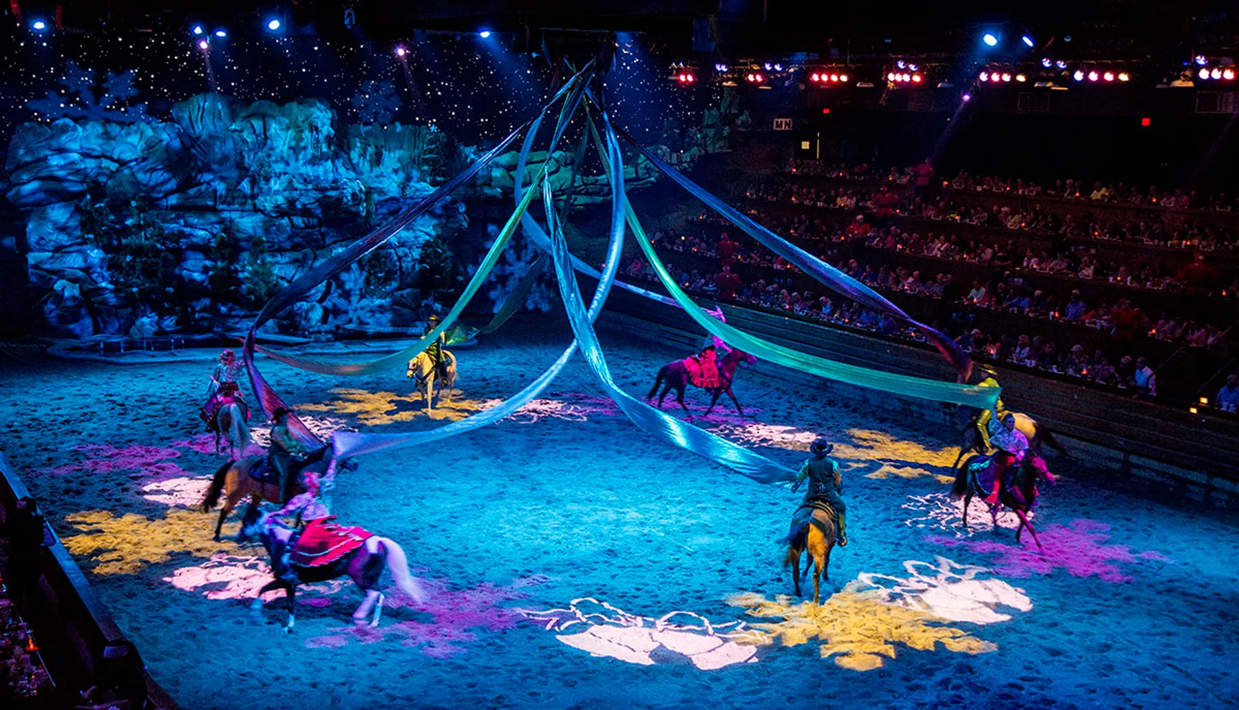 Riders on horseback perform in an equestrian-themed show before an audience in an arena illuminated with colorful lighting and decorated with theatrical scenery.