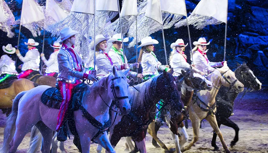 A group of riders in white outfits and cowboy hats are parading on horses under artificial snow, carrying large white umbrellas as part of a festive event or show.