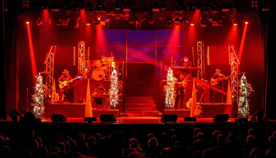 A band performs on a festively decorated stage with red lighting and Christmas trees as the audience watches.