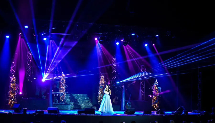 A person dressed in a long dress stands onstage, bathed in beams of blue stage lighting, surrounded by festive, illuminated Christmas trees, suggesting a holiday performance or concert.