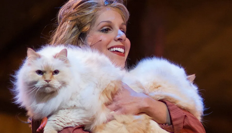 A smiling woman is holding a large, fluffy white cat with an intense gaze.