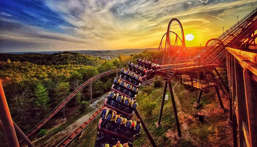 A roller coaster train ascends a track against a dramatic sunset sky, offering a picturesque view of the surrounding landscape.