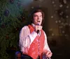 A person is performing on stage with a microphone in hand against a festively decorated backdrop with a Christmas tree and a large gift-wrapped box