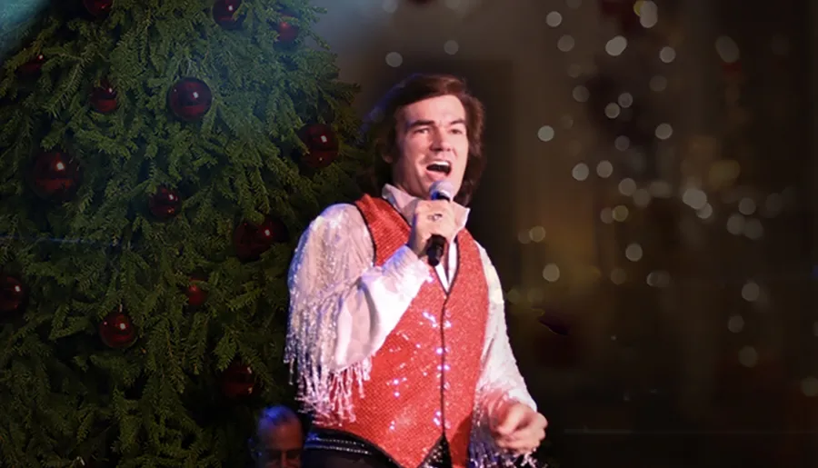 A person is performing on stage with a microphone in hand, wearing a red sequined vest, with a Christmas tree in the background suggesting a holiday theme.