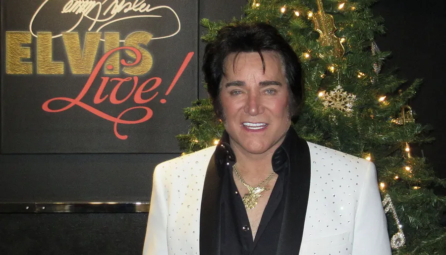 A person resembling Elvis Presley stands in front of a Christmas tree with a backdrop that reads Elvis Live!