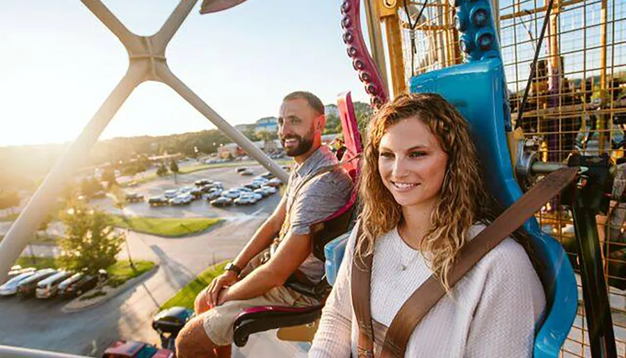 A smiling woman and a man enjoy a ride on a brightly colored Ferris wheel, with a view of the surrounding area basked in the glow of the setting sun.