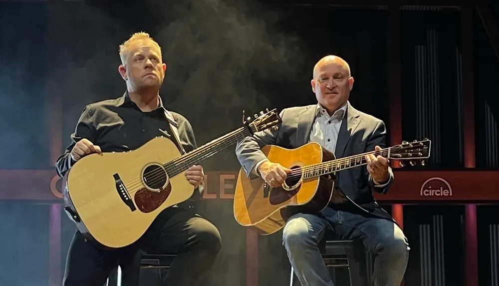 Two musicians are seated on stage playing acoustic guitars with a smoky backdrop highlighted by soft lighting