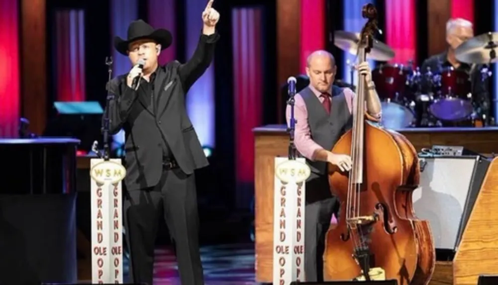 A performer in a cowboy hat is singing on stage at the Grand Ole Opry with a microphone stand labeled WSM while another musician plays an upright bass and a drummer is visible in the background