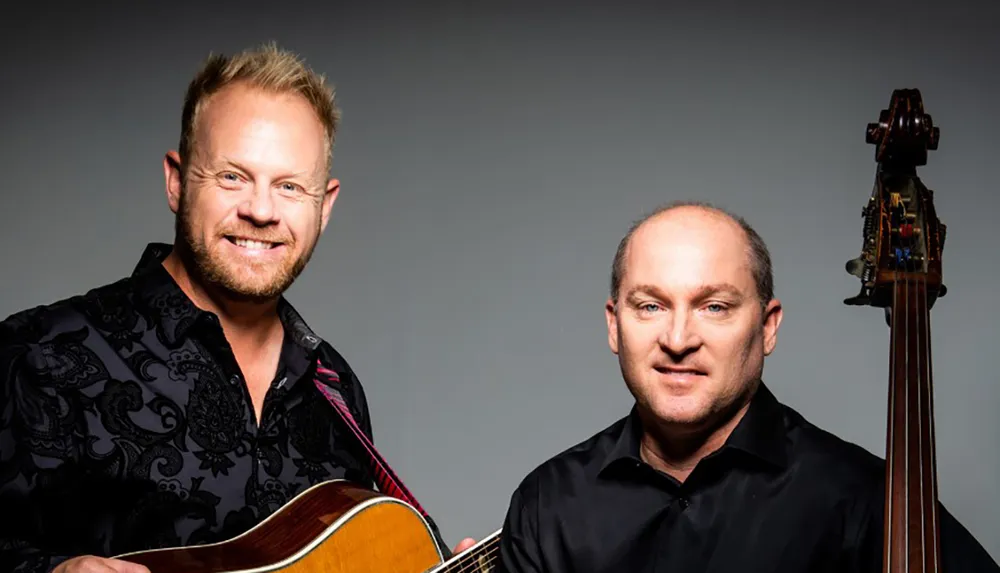 Two smiling men one holding a guitar and the other standing next to an upright bass pose for a portrait against a gray background