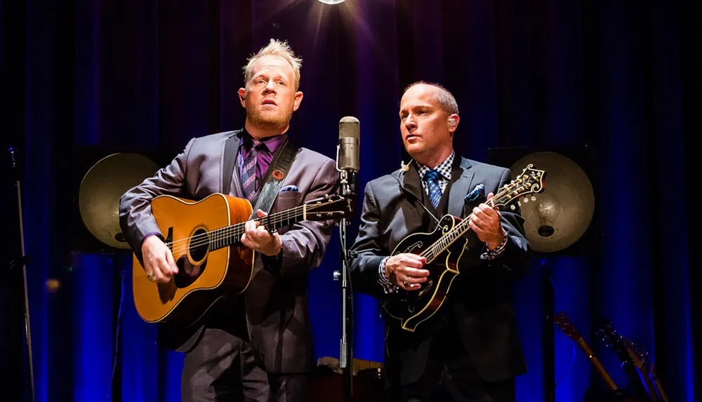 Two musicians one playing an acoustic guitar and the other a mandolin perform together on stage in front of a microphone