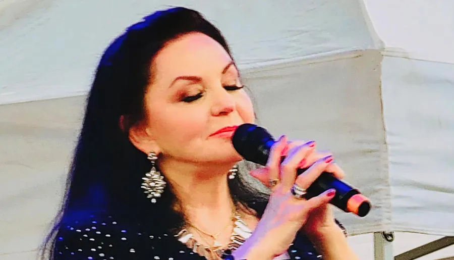 A woman with dark hair and elegant earrings is singing into a microphone with her eyes closed, expressing emotion in her performance.