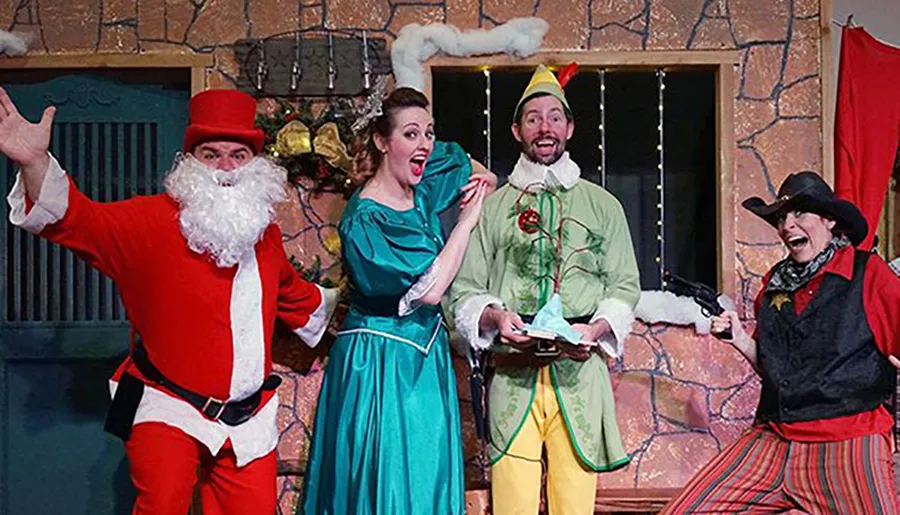 Four people are dressed in colorful and theatrical Christmas-themed costumes, expressing exaggerated emotions in what appears to be a festive play or performance.