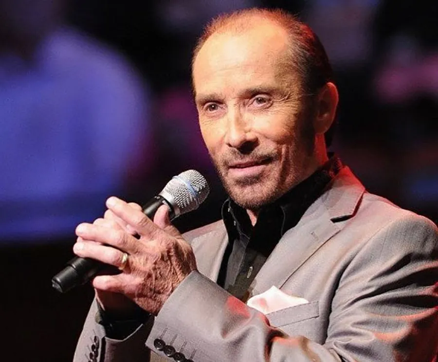 A man in a gray suit and tie is performing on stage with a microphone in hand.