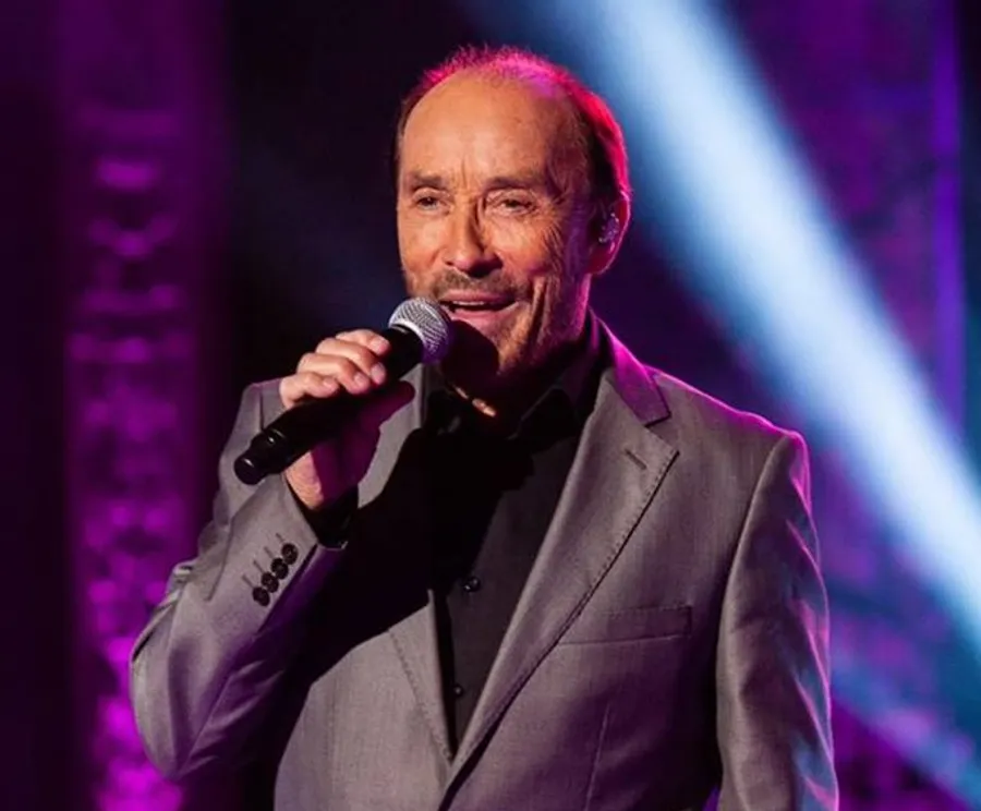 A smiling, elegantly dressed singer is performing on stage, illuminated by blue stage lights.