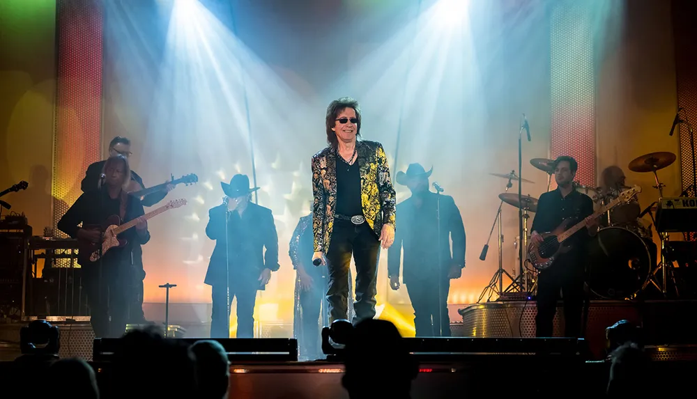 A band performs on stage with a charismatic lead singer in a distinctive jacket standing at the forefront under dramatic stage lighting