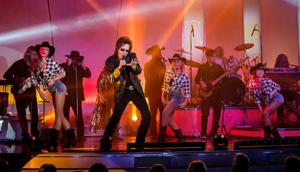 A performer sings passionately on stage flanked by musicians and dancers against a backdrop of warm stage lighting